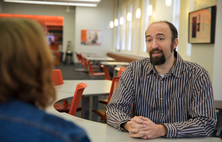 Speech communication professor Colin Hesse wears a brown button-up shirt with white horizontal stripes and his hands are folded on a curved white table in front of him. He is speaking to a person across the table whose head and shoulders are visible from behind. The person has shoulder length reddish-brown hair with light green roots and they wear a dark blue denim jacket. In the background, there is a room with more white tables and red rolling chairs.