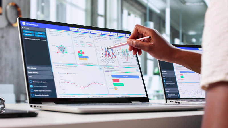 A computer screen shows charts, graphs and data visualizations. A hand holding a stylus points at the screen.