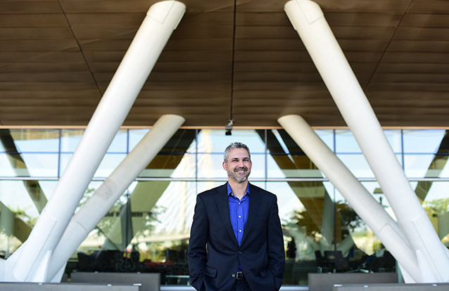 Eric Vela walks in front of a building with a glass front and white pillars extending diagonally from the ground to a large wood awning.