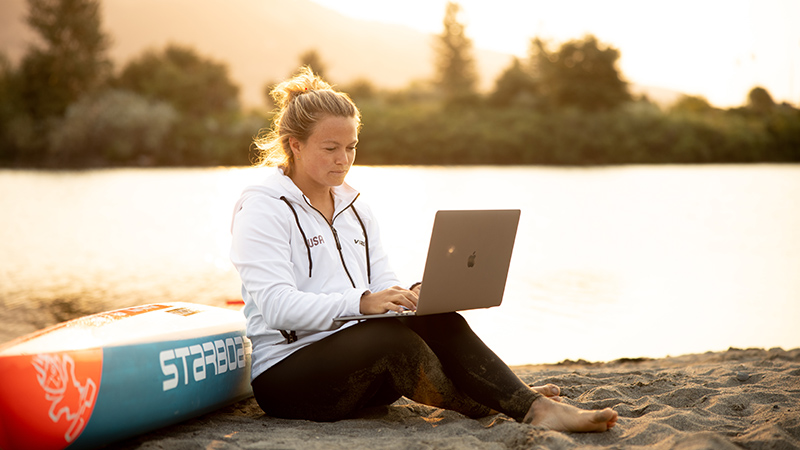 A barefoot woman sits on the beach sand next to a paddleboard while typing on a laptop that is resting on her knees. The sun reflects off the lake water behind her.