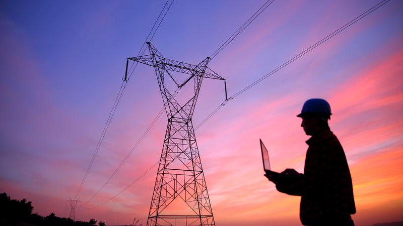 An energy grid worker stands outside near an electrical tower while holding a laptop computer.