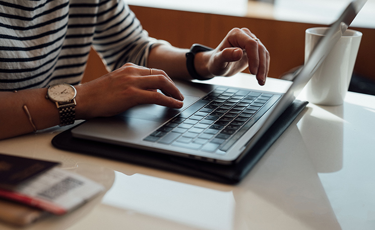 A person sits at a cafe table and works on a laptop, with a notebook at their right and a coffee mug to their left. Online enrollment services