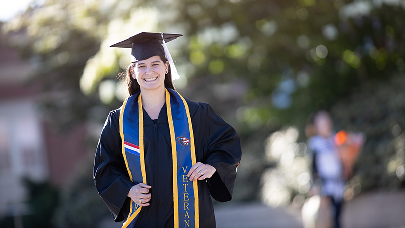 Hope Nelson, an Oregon State University alum, stands outside in commencement cap and gown with a blue sash that says veteran.