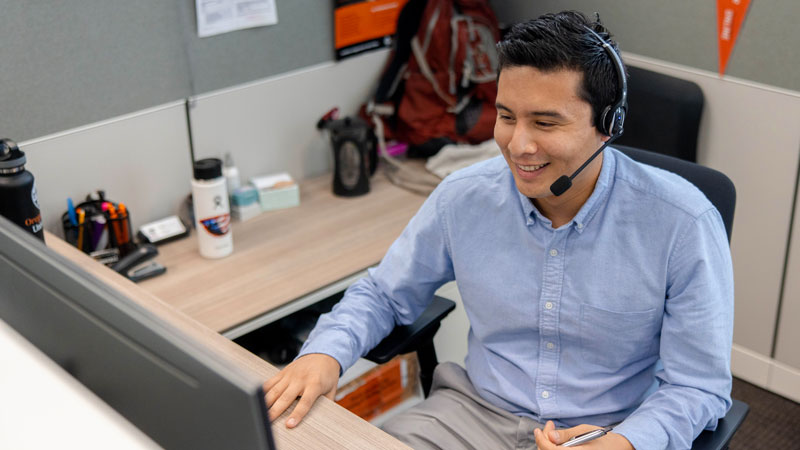 A student representative sits at a desk wearing a headset. A computer, phone and cups of water are visible.