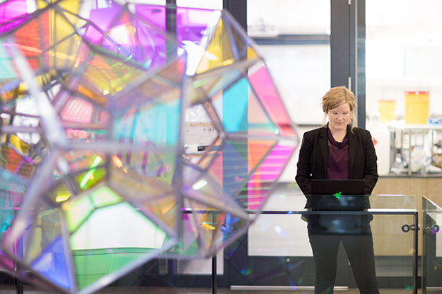 Ecampus engineering management student Ingrid Scheel stands on a balcony with a glass barrier. There is a hanging multi-colored glass sculpture near her.