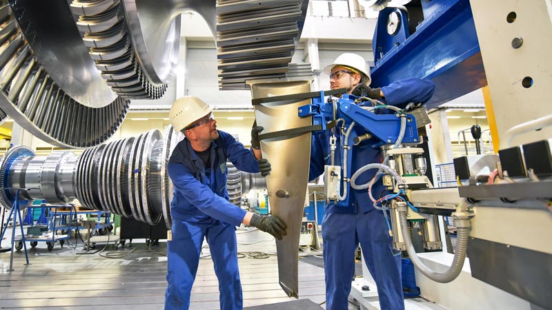 Two engineers wearing blue coveralls install a blade on a mechanical turbine.