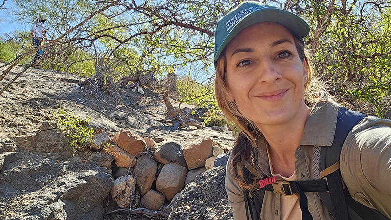 An outdoor selfie of Oregon State University student Melissa Croteau, kneeling in front of a dry, rocky hill adorned with small green shrubs and trees.