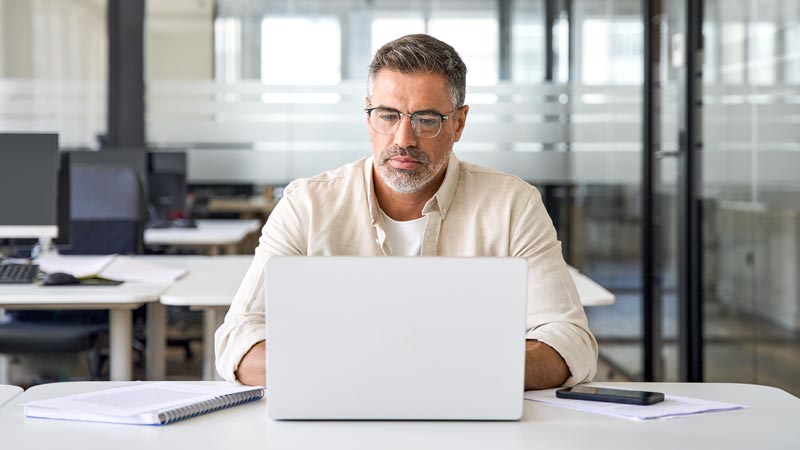 A man sits at a table while working on a laptop computer.