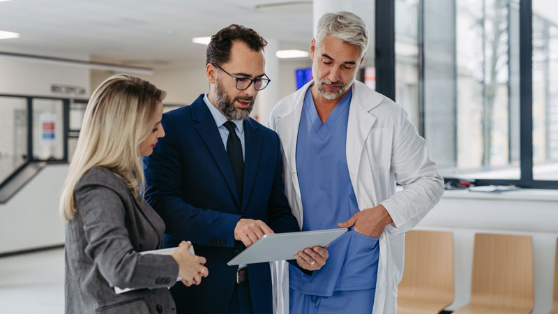 Three professionals – a woman in a gray blazer, a man in a blue suit, and a man in blue scrubs and a lab coat – converse while looking at a tablet.