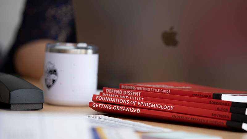 A close-up image of a stack of orange textbooks on a desk. The books are next to a white coffee mug with an Oregon State University logo on it and a laptop.