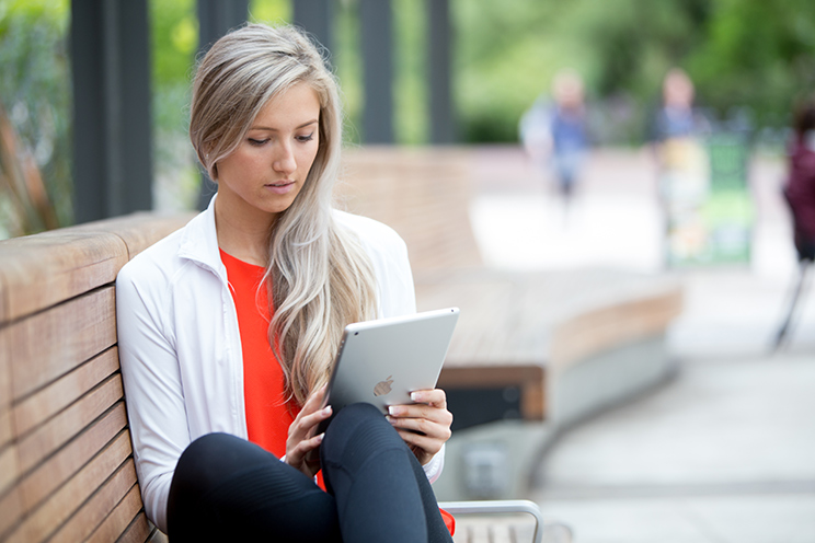 A woman sits on a bench outside and works on a smart tablet, completing an online application.