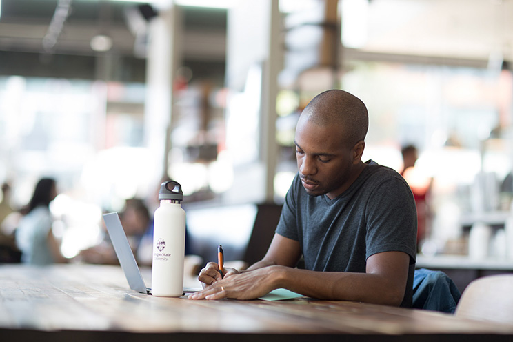 A man sits at a table in a bright cafe while writing on a notepad. A white water bottle and open laptop are near him.
