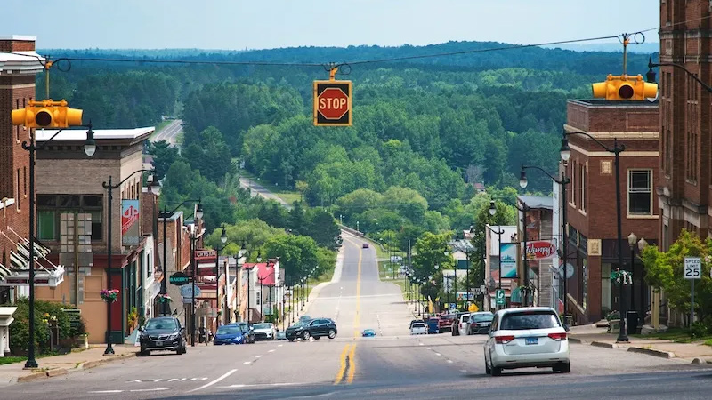 A view of a small town, looking down a gently sloping street lined with buildings on either side, leading into a lush, green wooded area.