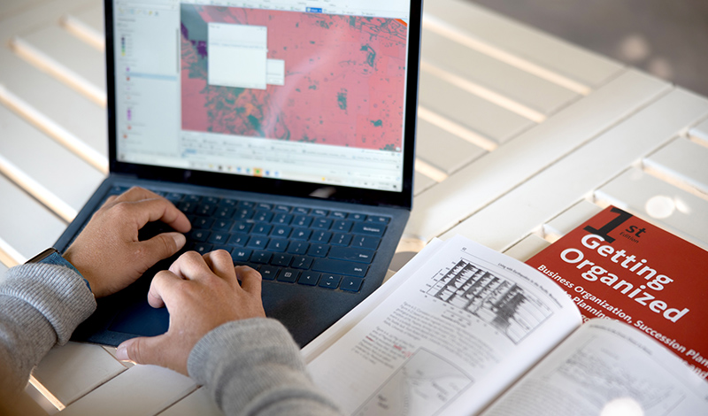 A person working on a laptop with a geographic map on the screen, alongside an open textbook titled "Getting Organized."