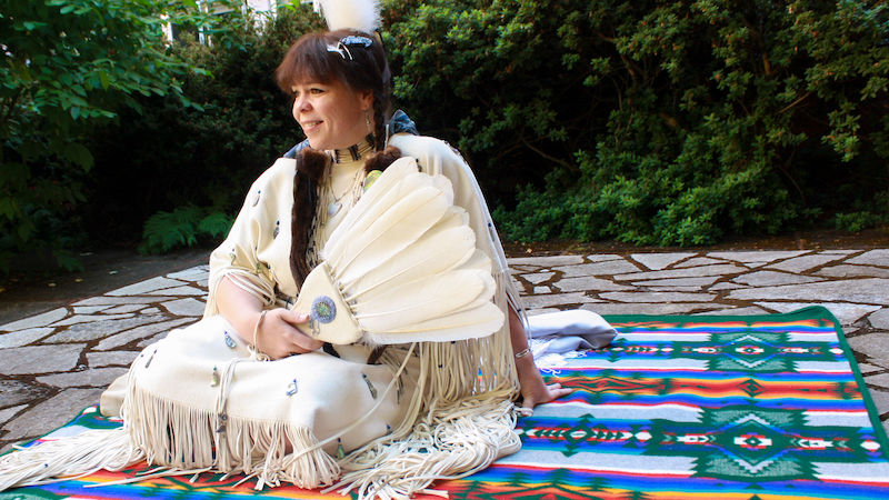 Samantha Chisholm Hatfield sits on a traditional tribal blanket in a traditional garment, holding a fan made of feathers.