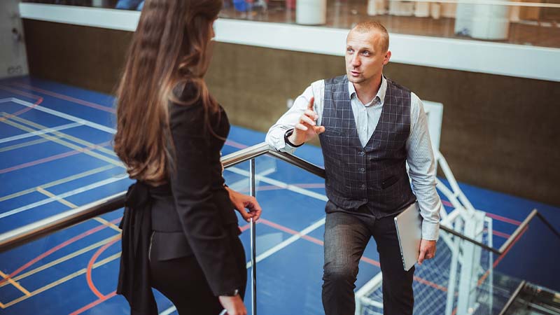 Two people stand on stairs within a sports facility. One person talks while holding a clipboard.