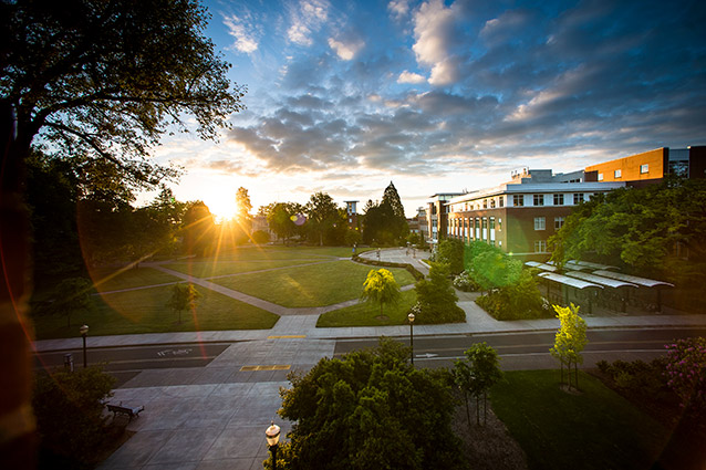 The Valley Library quad at Oregon State University is photographed from a high vantage point. There are clouds in the sky and the sun is shining brightly through trees at the edge of the quad.