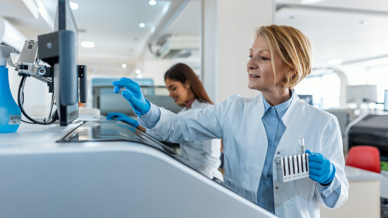 A person in a white lab coat and blue gloves hold test tubes while standing in front of a machine. Another person in a white coat looks at that machine.
