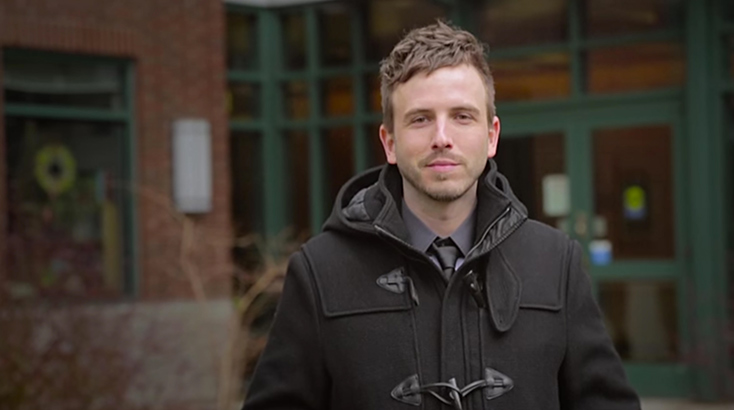 Justin Zweifel wears a black coat and stands in front of a red brick building with dark green trim.