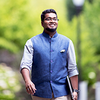 Oregon State Ecampus graduate Patric Papabathini walks forward with a smile on his face and a laptop carried under his left arm. He is wearing a half-sleeve, light blue button-up shirt under a dark blue Nehru collar vest with a handkerchief in the breast pocket.