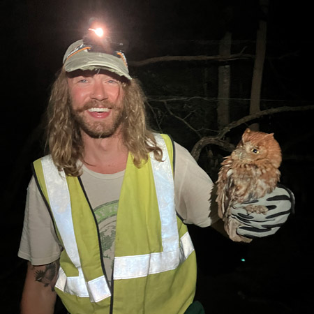 This picture depicts Bill, a white man with long blond hair, wearing a high visibility vest, a ball cap, and a head light because this picture was taken at night. In his gloved hands, he holds a baby owl. Bill is smiling widely.