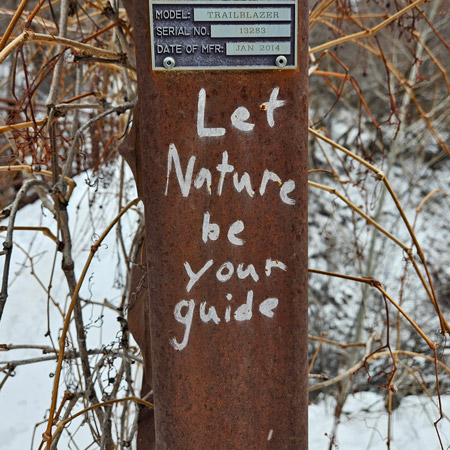This photo depicts a close up of pole of a bridge. It’s brown, rusted, with a silver plaque that give the hiker information about how much weight the bridge can hold, the serial number, etc.. On the brown, rusted part of the pole, someone has written in bright white lettering, “Let nature be your guide.” The white lettering pops out from the rusted pole and connects with the snowy background.