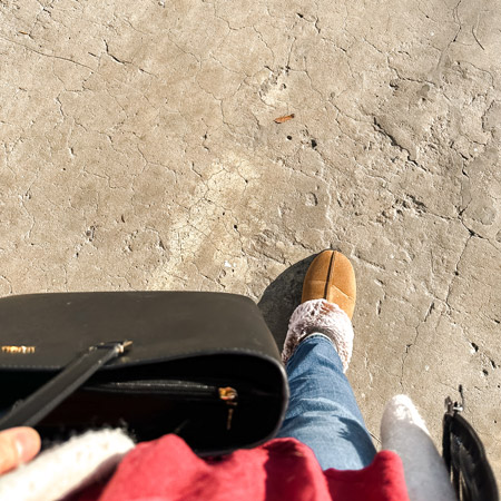 This picture shows us Veronica B’s view looking down at her feet as she walks across cement. We see her right leg in denim, her right foot in a light brown slipper, and she’s holding a black bag close to her red shirt.